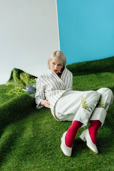 Bela menina loira elegante posando na grama artificial com samambaia e flores — Fotografia de Stock