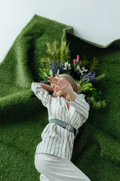 Stylish girl covering face with hand while lying on artificial grass with fern and flowers — Stock Photo