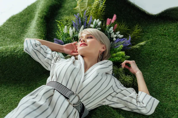Schöne stilvolle Mädchen mit geschlossenen Augen posiert auf Kunstrasen mit Farn und Blumen — Stockfoto