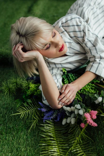 Belle fille blonde élégante avec les yeux fermés couché sur gazon artificiel avec fougère et fleurs — Photo de stock