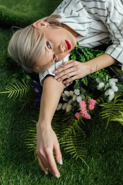 Красива стильна дівчина дивиться на камеру, лежачи на штучній траві з папороті і квітами — стокове фото