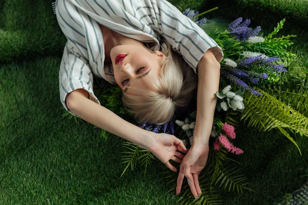 Visão de alto ângulo da bela menina elegante com os olhos fechados deitado na grama artificial com samambaia e flores — Fotografia de Stock