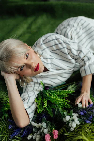 Bela menina elegante olhando para a câmera enquanto deitado na grama artificial com samambaia e flores — Fotografia de Stock