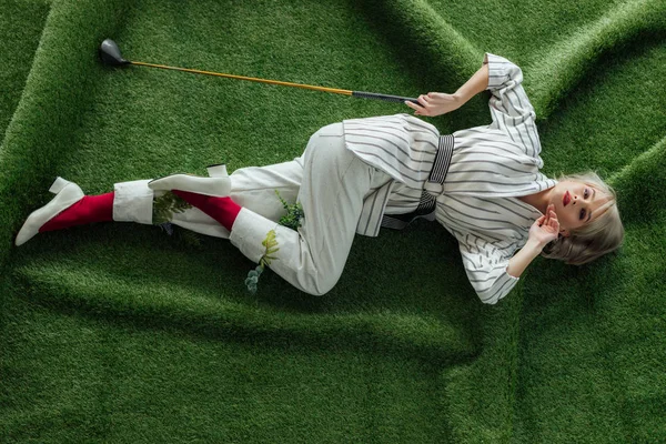 Hermosa chica elegante tumbado con club de golf en césped artificial y mirando a la cámara - foto de stock