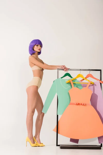 Belle fille en sous-vêtements avec les cheveux violets en regardant la caméra tout en posant près de rack avec des vêtements en papier coloré sur blanc — Photo de stock