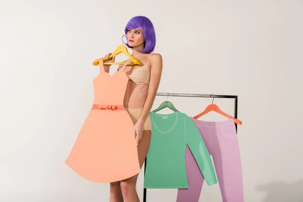 Attractive girl with purple hair holding paper dress and posing near rack with colorful clothes on white — Stock Photo