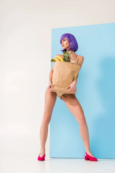 Hermosa chica con el pelo morado posando mientras sostiene bolsa de papel con frutas en azul y blanco - foto de stock