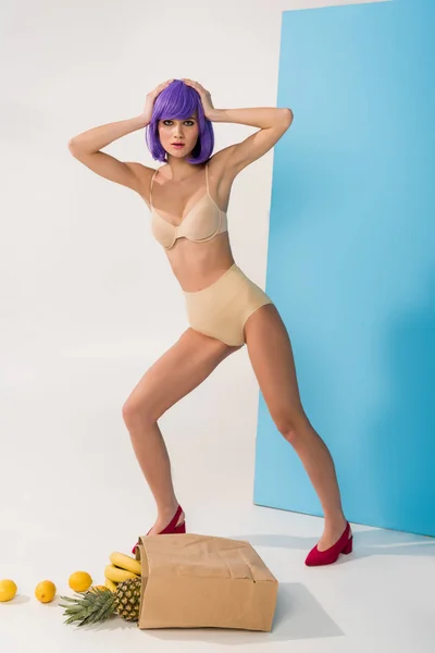 Hermosa chica con el pelo morado mirando a la cámara mientras posa cerca de bolsa de papel con frutas tropicales en azul y blanco - foto de stock