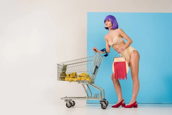 Hermosa chica con el pelo morado posando cerca del carrito de la compra con frutas tropicales en azul y blanco con espacio de copia - foto de stock
