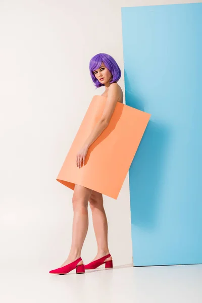 Menina atraente com cabelo roxo coberto de folha de papel de coral olhando para a câmera enquanto posando em azul e branco — Fotografia de Stock