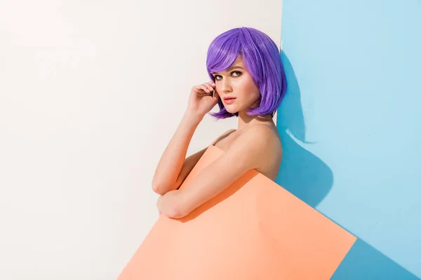 Belle fille aux cheveux violets recouverts de feuille de papier corail regardant la caméra tout en posant sur bleu et blanc — Photo de stock