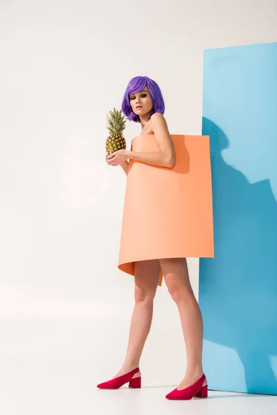 Belle jeune femme aux cheveux violets recouverts de feuille de papier corail tenant l'ananas sur bleu et blanc — Photo de stock