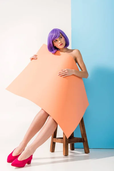 Beautiful young woman with purple hair covered in coral paper sheet sitting on chair while posing on blue and white — Stock Photo