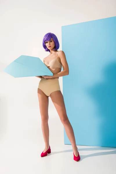 Hermosa mujer joven con el pelo púrpura mirando a la cámara mientras posa con el avión de papel en azul y blanco - foto de stock