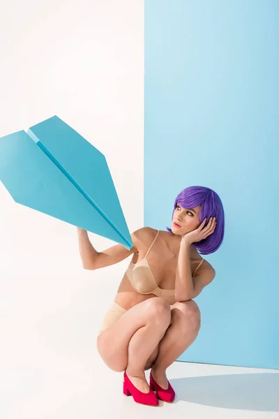 Belle fille aux cheveux violets tenant avion en papier tout en posant sur bleu et blanc — Photo de stock