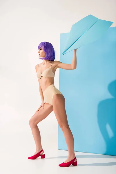 Schöne Mädchen mit lila Haaren hält Papierflieger, während sie auf blau-weiß posiert — Stockfoto