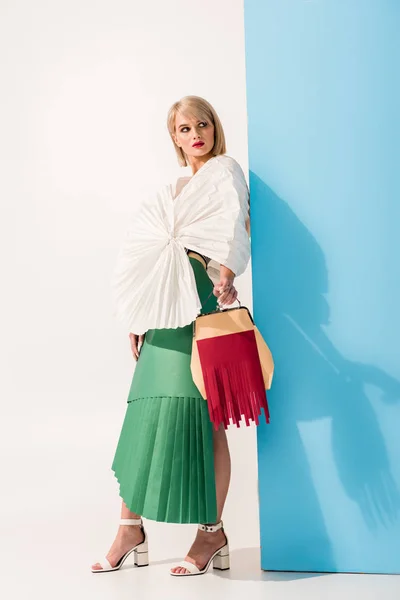 Hermosa chica con estilo en ropa de papel sosteniendo bolsa de papel y posando en azul y blanco - foto de stock