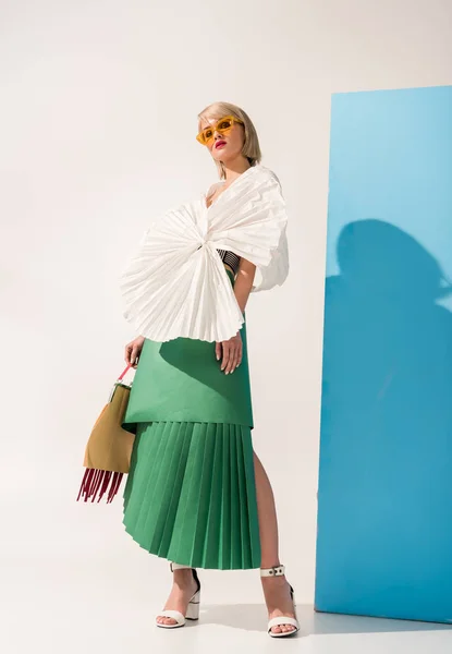 Hermosa chica con estilo en gafas de sol y ropa de papel mirando a la cámara mientras posan en azul y blanco - foto de stock