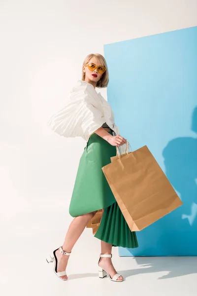 Красива стильна дівчина в сонцезахисних окулярах і паперовому одязі позує з сумками на синьому і білому — стокове фото