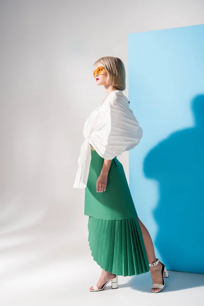 Belle fille élégante dans des lunettes de soleil et des vêtements en papier posant sur bleu et gris — Photo de stock