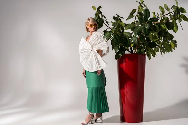 Bela menina elegante em óculos de sol e roupas de papel posando perto da planta no cinza com espaço de cópia — Fotografia de Stock