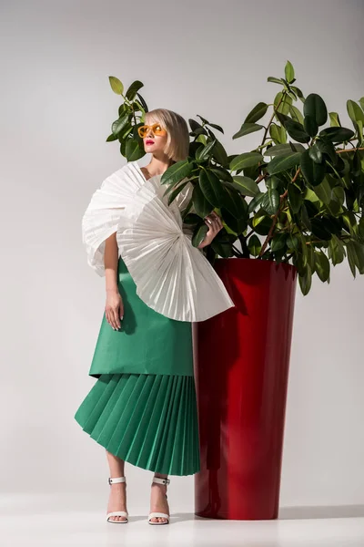 Hermosa chica con estilo en gafas de sol y ropa de papel posando cerca de la planta en gris - foto de stock