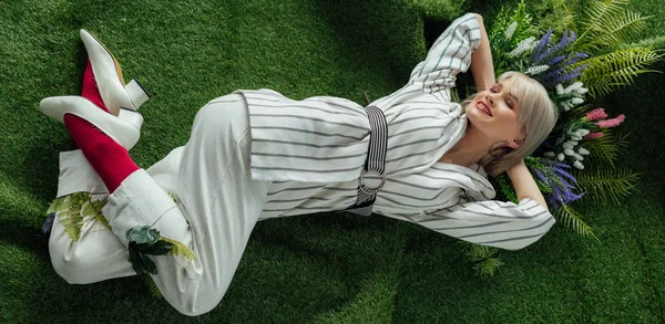 Panoramic shot of beautiful stylish girl smiling while lying on artificial grass with flowers — Stock Photo