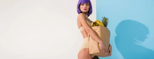 Plano panorámico de hermosa chica con pelo púrpura celebración bolsa de papel con frutas en azul y blanco - foto de stock
