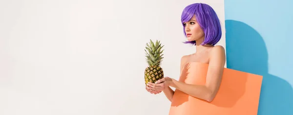Menina bonita com cabelo roxo coberto de folha de papel de coral segurando abacaxi enquanto posando em azul e branco — Fotografia de Stock