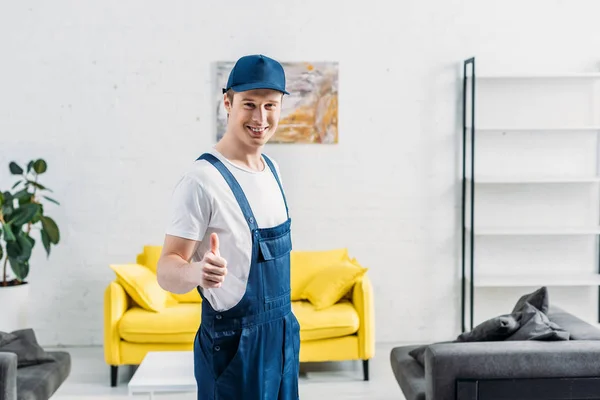 Lächelnder Mover in Uniform blickt in die Kamera und zeigt Daumenhoch-Zeichen in Wohnung — Stockfoto