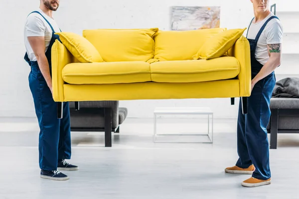 Vue recadrée de deux déménageurs en uniforme transportant canapé dans l'appartement — Photo de stock