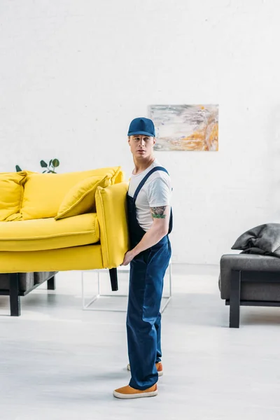 Handsome mover in uniform transporting couch in apartment — Stock Photo