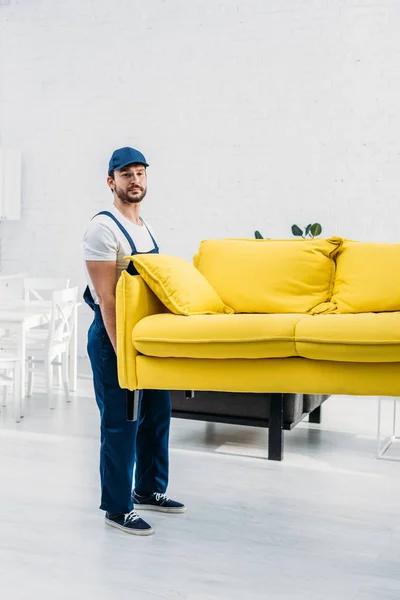 Handsome mover in uniform transporting couch in apartment — Stock Photo