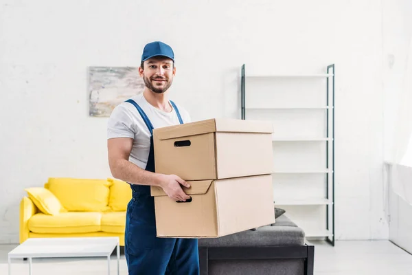 Schöner Mover in Uniform, der in die Kamera schaut, während er Kartons in einer Wohnung mit Kopierraum trägt — Stockfoto