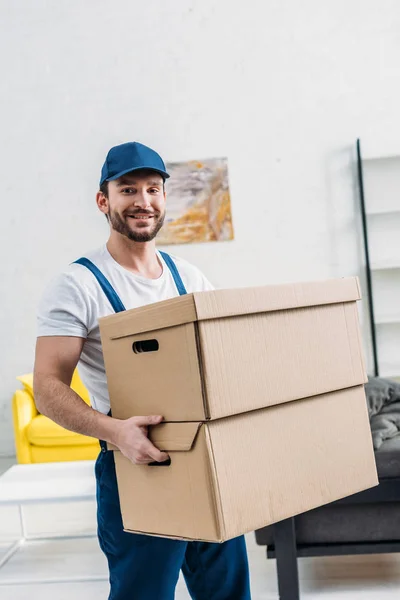 Guapo mover en uniforme mirando a la cámara mientras lleva cajas de cartón en apartamento - foto de stock