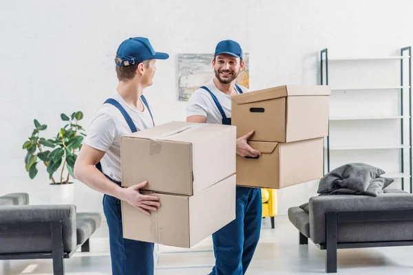 Two movers in uniform looking at each other while carrying cardboard boxes in apartment — Stock Photo