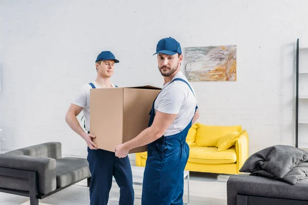 Two handsome movers in uniform transporting cardboard box in apartment — Stock Photo