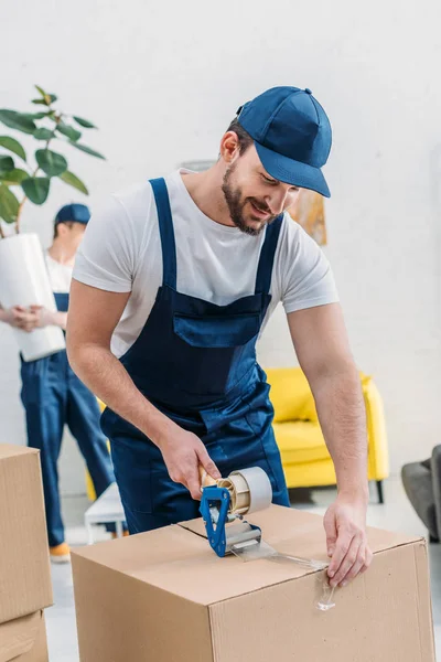 Handsome mover in uniform wrapping cardboard box with scotch tape in apartment — Stock Photo