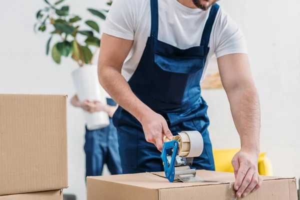 Cropped view of mover wrapping cardboard box with scotch tape in apartment — Stock Photo