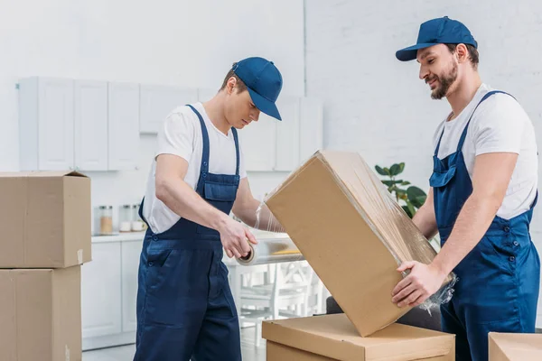 Handsome movers wrapping cardboard box with stretch film in apartment — Stock Photo