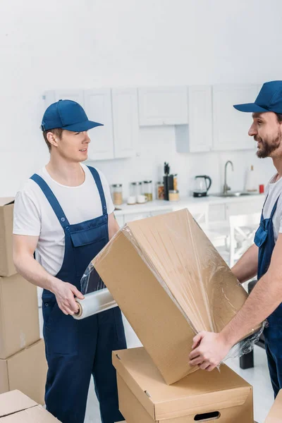 Movers in uniform wrapping cardboard box with stretch film in apartment — Stock Photo