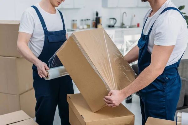 Partial view of movers wrapping cardboard box with stretch film in apartment — Stock Photo