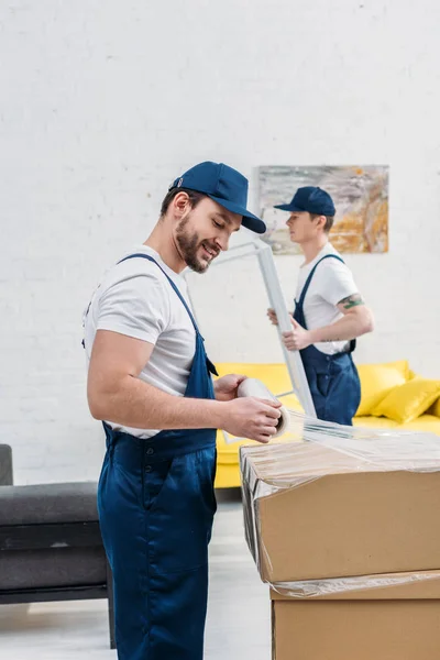 Handsome mover in uniform wrapping cardboard box with stretch film in apartment — Stock Photo