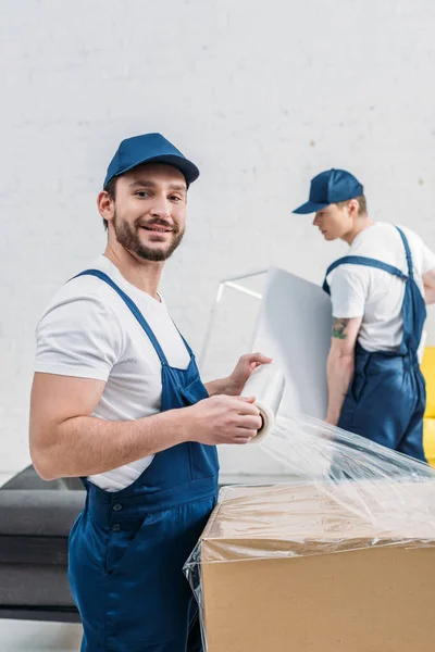 Handsome mover in uniform looking at camera while wrapping cardboard box with stretch film in apartment — Stock Photo