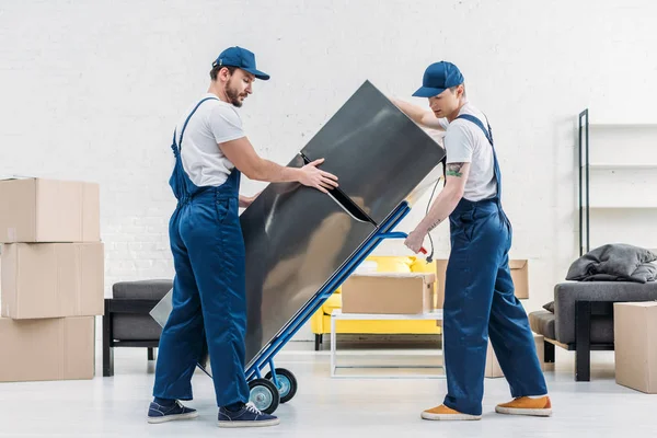 Two movers using hand truck while transporting refrigerator in living room — Stock Photo