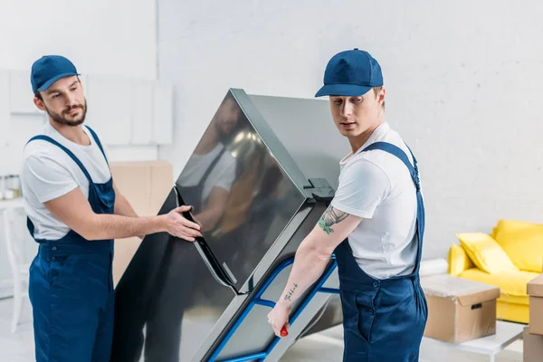 Two movers in uniform using hand truck while transporting refrigerator in apartment — Stock Photo