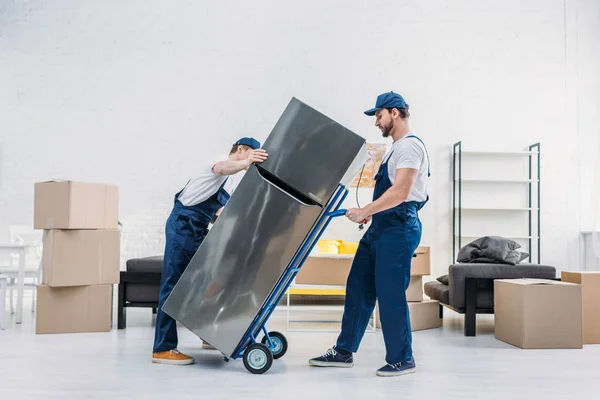 Two movers in uniform using hand truck while transporting refrigerator in apartment — Stock Photo