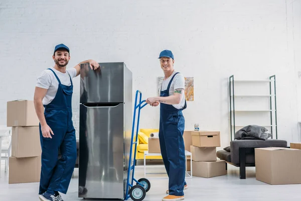 Two smiling movers using hand truck while transporting refrigerator in apartment — Stock Photo