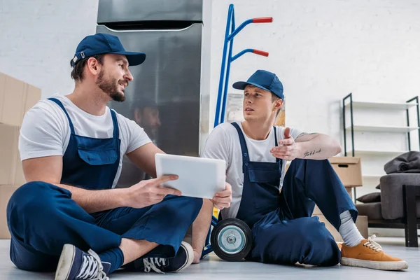 Two movers in uniform using digital tablet while sitting near hand truck and refrigerator in apartment — Stock Photo