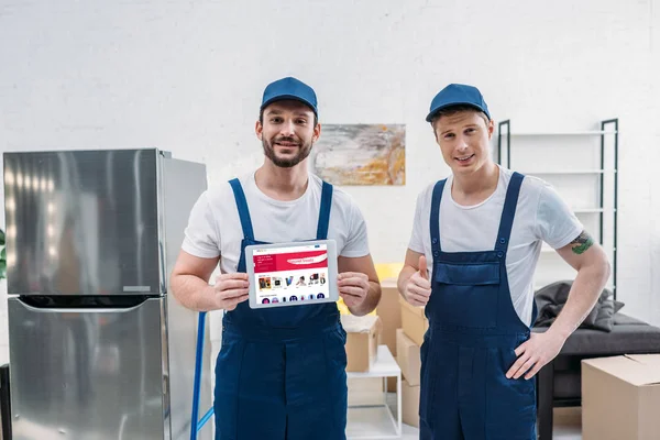 Two movers presenting digital tablet with ebay app on screen and showing thumb up sign in apartment — Stock Photo